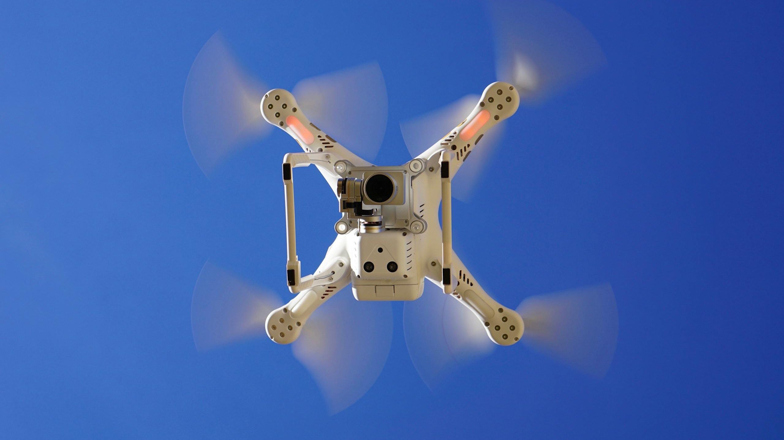 Aerial view of white drone flying with rotating blades against a clear blue sky, capturing video footage.